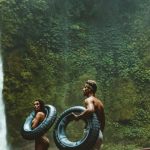 Adventures - Two Person Carrying Black Inflatable Pool Float on Brown Wooden Bridge Near Waterfalls